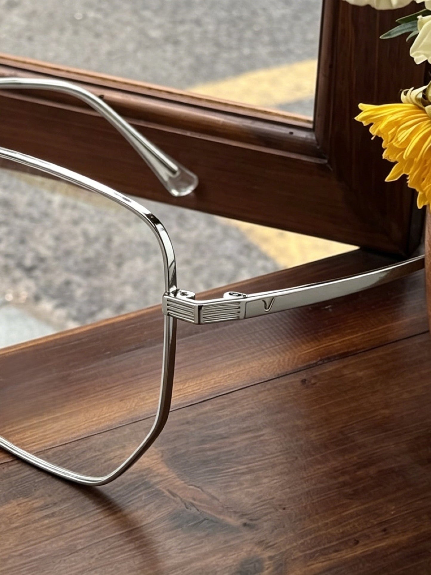 :Close-up shot of silver mens wire frame glasses hinge detail, textured metal temple with V logo on wooden window surface, lightweight thin wire metal frame men prescription eyewear detail photography