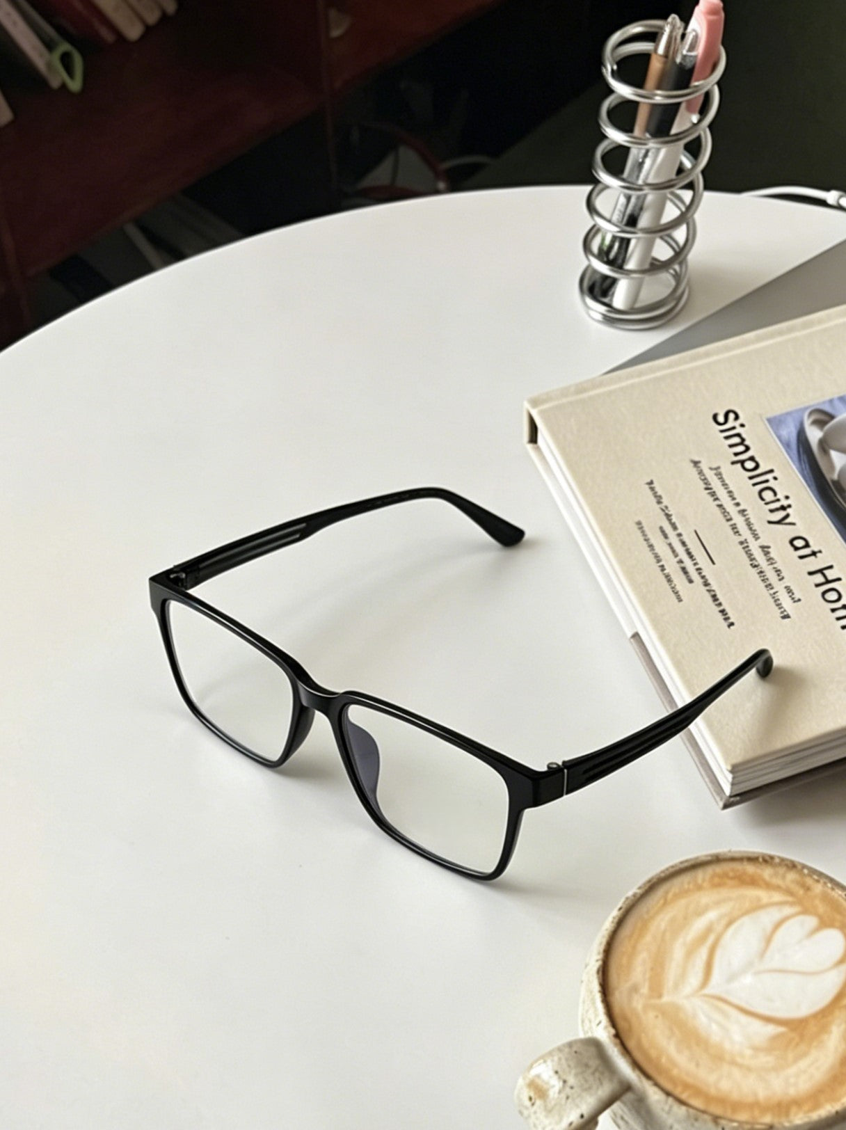 Men's black rectangle glasses flat lay on white round desk with latte coffee and hardcover notebook, minimalist office home workspace eyewear styling black rectangular acetate frame