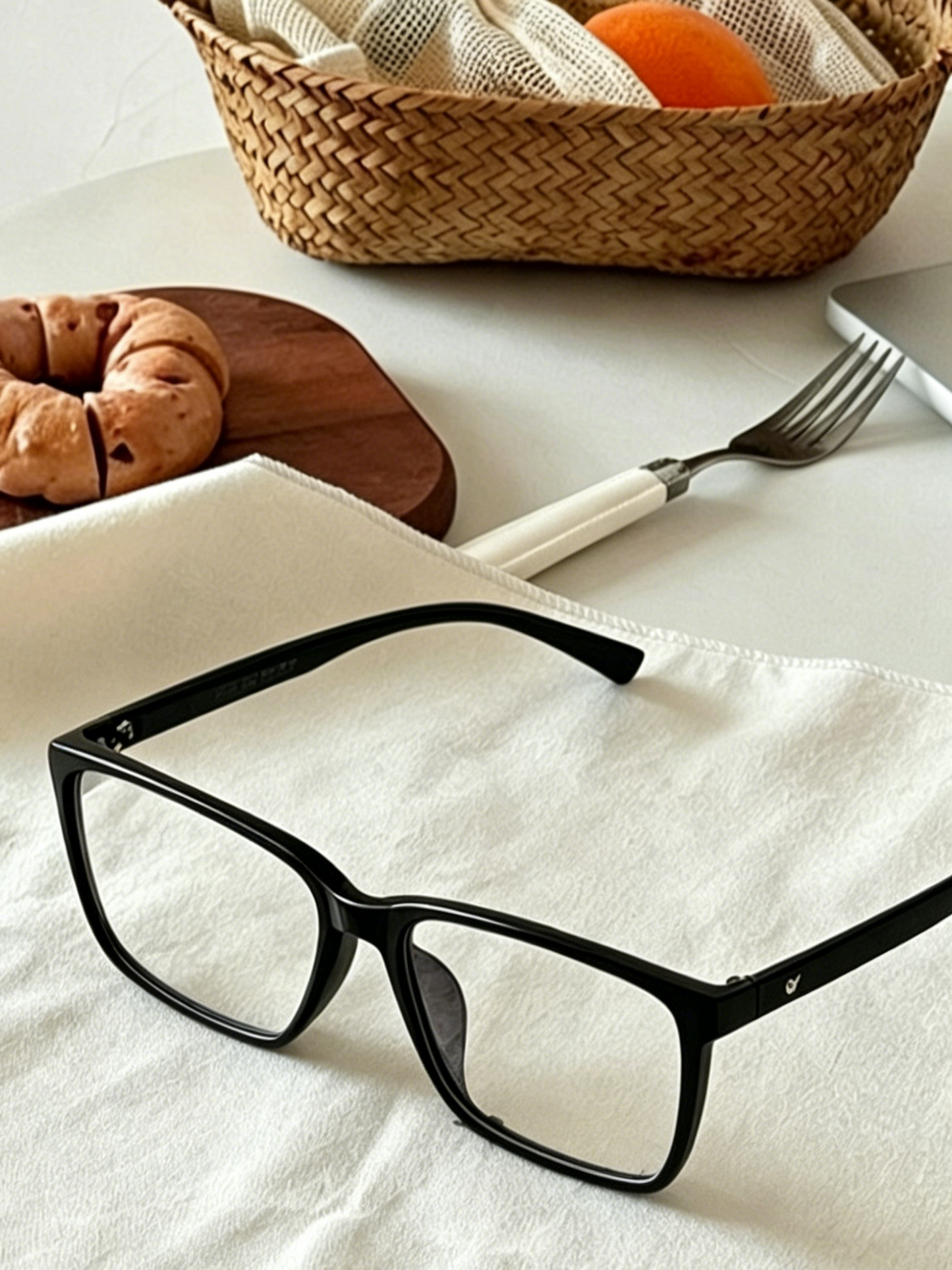 Close-up shot of black rectangle glasses frames on white linen napkin, breakfast table with bagel and woven basket, classic rectangular women men eyewear detail photography