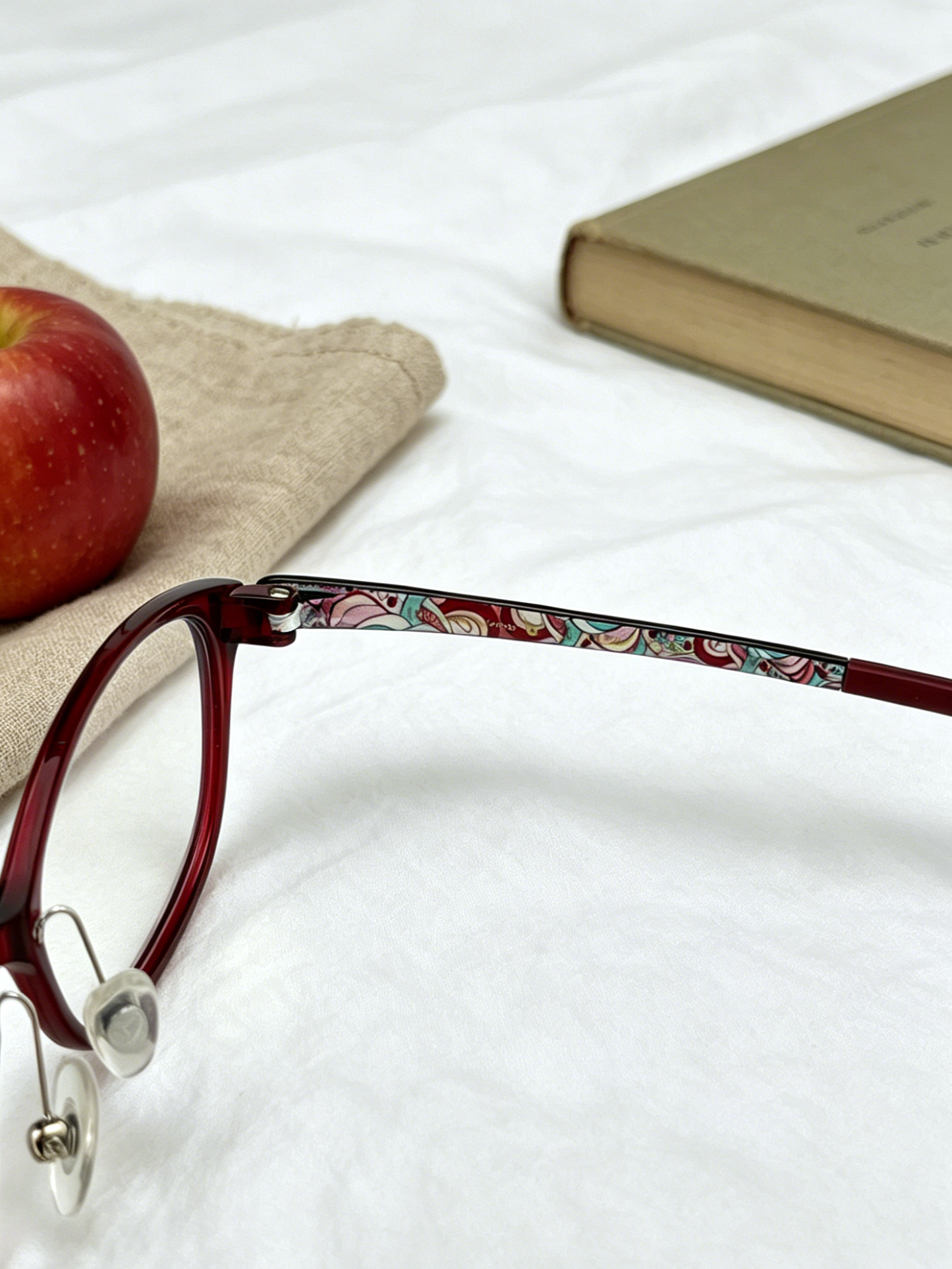 Close-up shot of burgundy cat eye glasses temple with colorful floral print detail, linen cloth and red apple background, thin acetate cat eye women eyewear texture photography