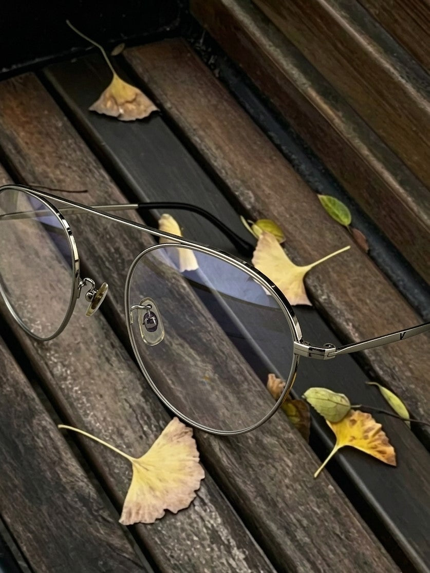 Close-up shot of aviator metal glasses on wooden bench surrounded by yellow ginkgo leaves, thin silver metal double bridge aviator eyeglass frame autumn detail photography