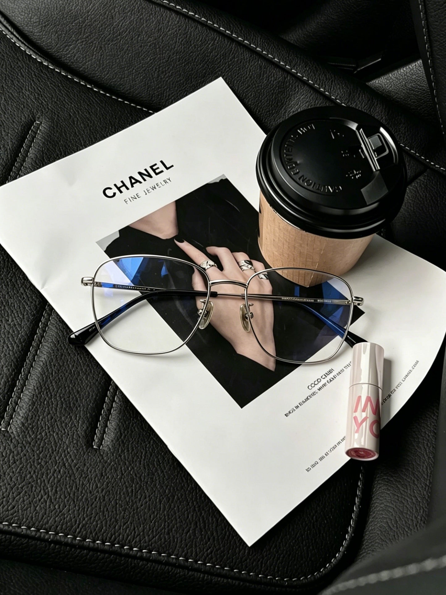 Thin metal glasses frames flat lay on black leather car seat with fashion magazine, takeaway coffee and lip gloss, minimalist car interior aesthetic eyewear styling silver metal frame