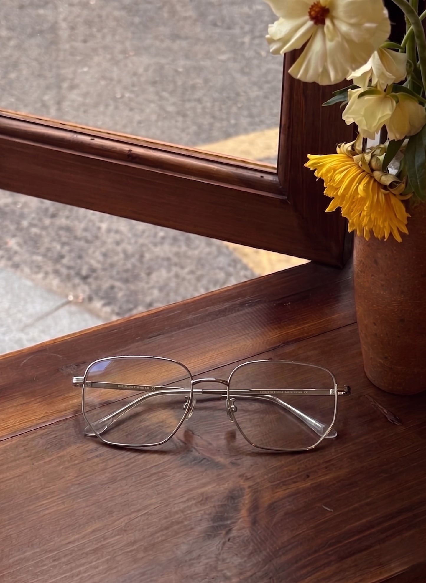 Silver mens wire frame glasses flat lay on vintage wooden window sill with yellow and white flowers, warm neutral cafe aesthetic men eyewear styling thin hexagonal wire metal eyeglass frame