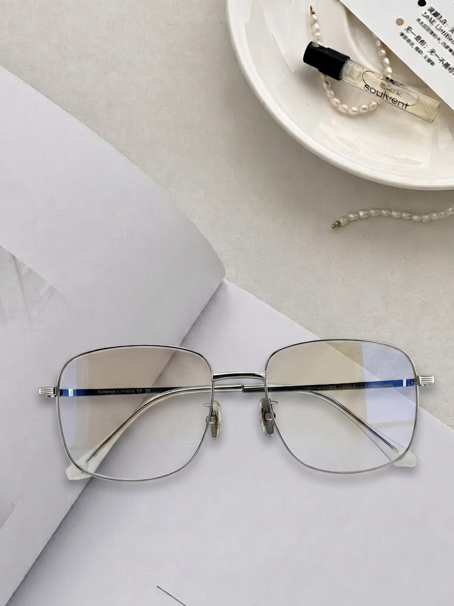 Silver oversized square glasses flat lay on cream paper with perfume bottle and pearl necklace, minimalist neutral desktop aesthetic thin metal square eyewear styling for daily office